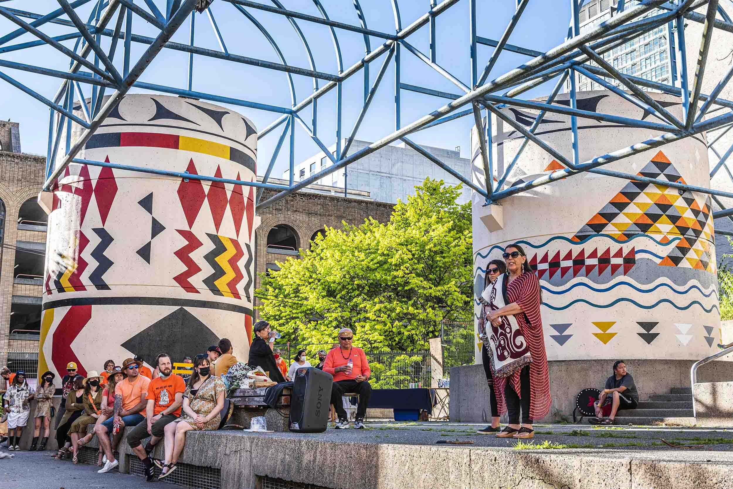 Debra sparrow speaking in front of audience at Cathedral Square in Downtown Vancouver. Two large cement pillars are in the background, they are painted with Coast Salish mural designs.
