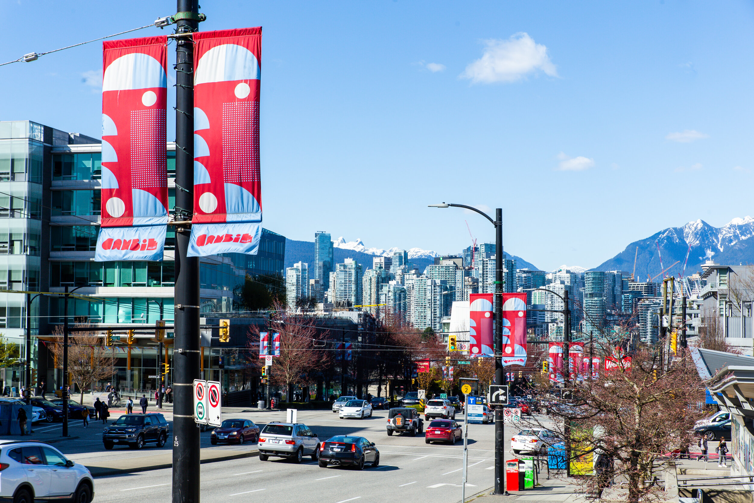 View from Cambie Village with cars on road looking North out onto the city of Vancouver.  Vibrant Cambie Village banners are dotted throughout in blue and red